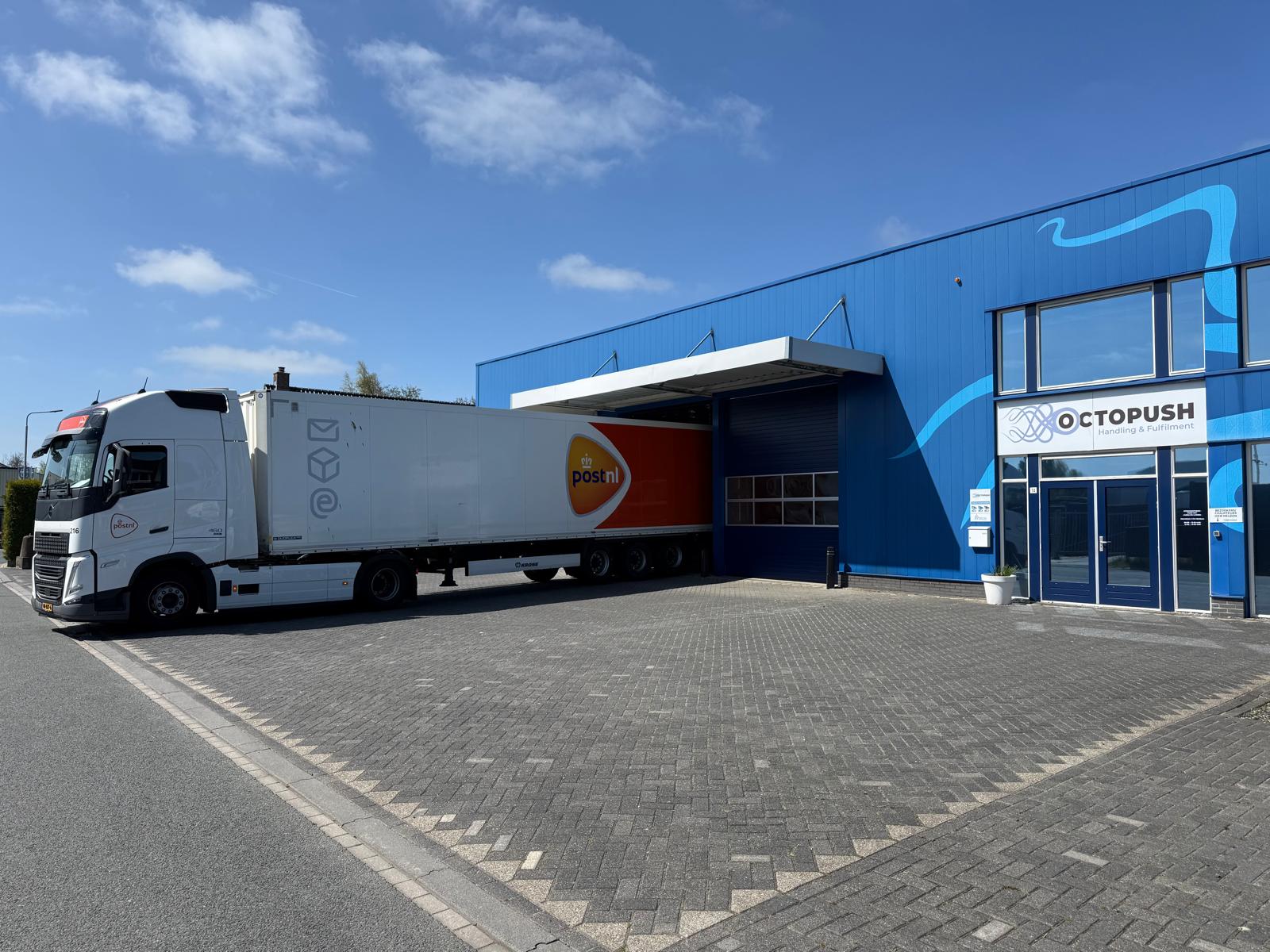 Voor een blauw gebouw met een Octopush-bord op de glazen deur staat een wit-oranje vrachtwagen van PostNL geparkeerd. De lucht is helder en zonnig.