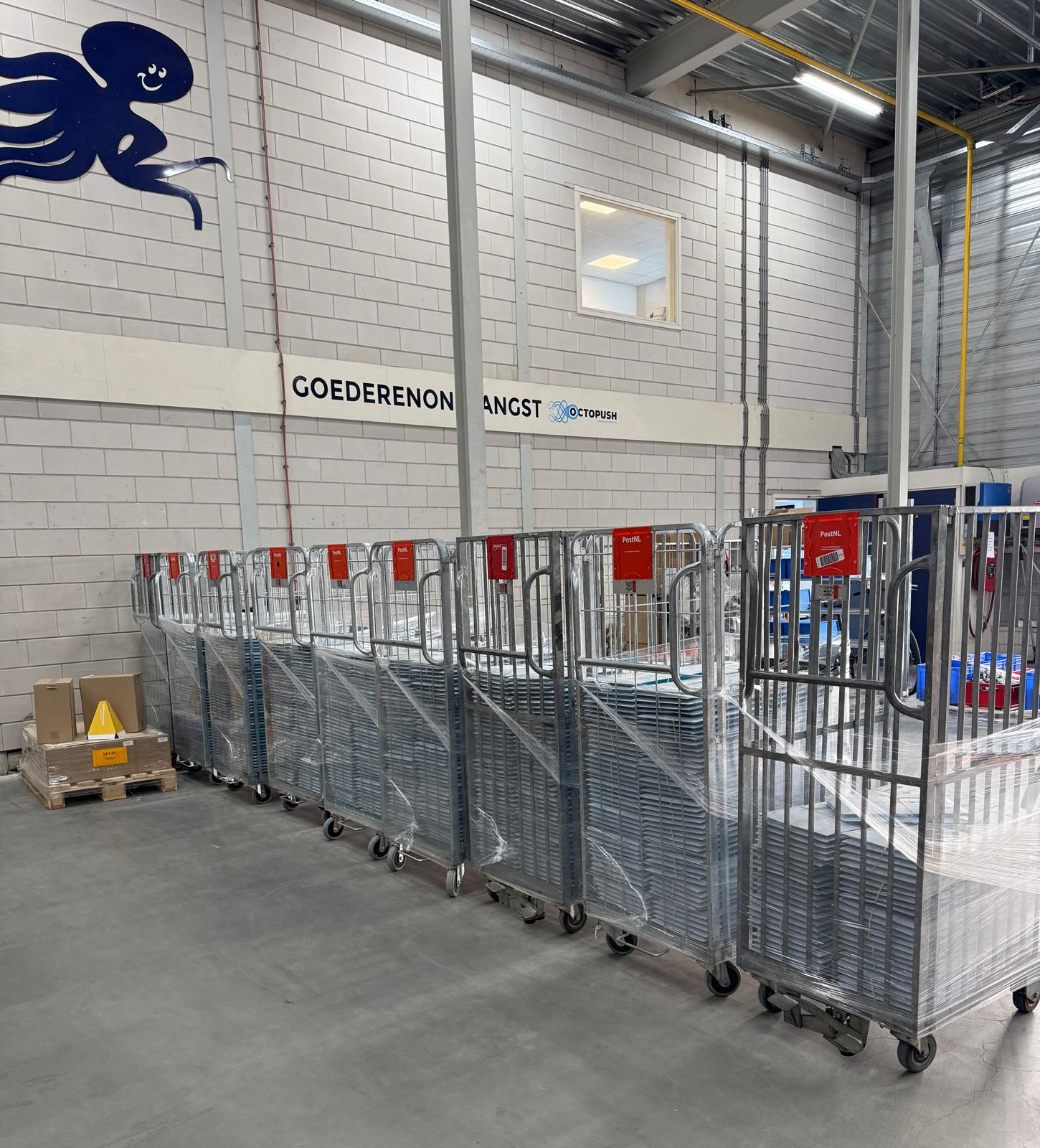 Magazijn met een rij metalen rolwagens, een paar kartonnen dozen op een pallet en een muur waarop ‘GOEDERENONTVANGST’ staat geschreven. Linksboven op de muur is een afbeelding van een blauwe octopus te zien. Een lichte, schone industriële omgeving.